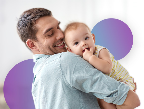 A father is holding his baby in his hands smiling at them whilst the baby is playing with their hands