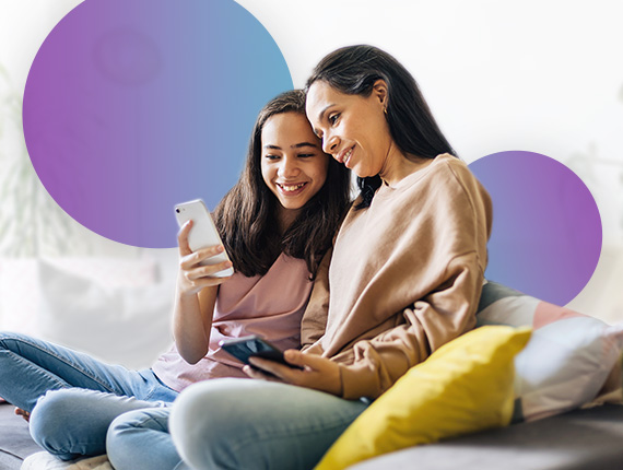 A mum and her teenage daughter sit together on the sofa looking at the daughter's mobile phone screen.