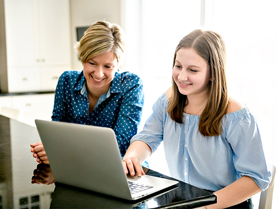 A mum and her teenager daughter both smiling as they read from a laptop at their dining table.