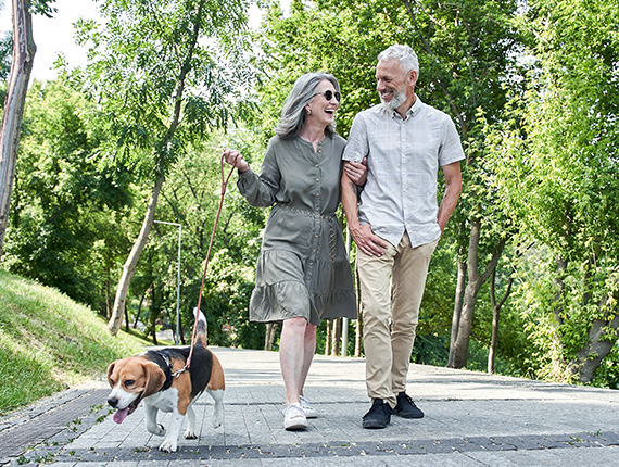 Retired couple smiling as they stroll down a tree-lined street, walking their dog